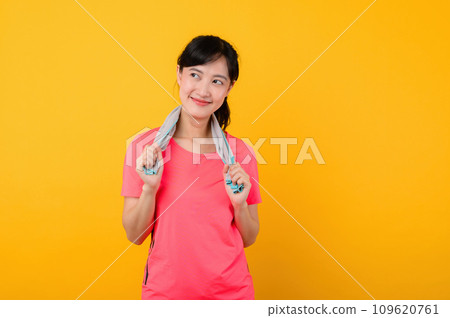 Portrait young asian sports fitness woman happy smile wearing pink sportswear and face towel doing exercise training workout against yellow studio background. wellbeing and healthy lifestyle concept. 109620761