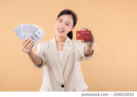 portrait of happy successful confident young asian business woman wearing white jacket holding cash money dollars and credit card standing over beige background. millionaire business, shopping concept 109620830