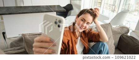 Portrait of beautiful, happy young female model, sitting on couch with smartphone, taking selfie on mobile phone, making cheerful smiling face 109621022