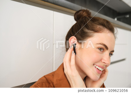 Close up portrait of girl enjoying listening to music in wireless earphones, smiling, wearing headphones, sitting at home 109621056