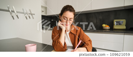 Portrait of young woman, student doing her homework, studying at home, sitting in kitchen, making notes, writing down information Portrait of young woman, student doing her homework, studying at home, sitting in kitchen, making notes, writing down information 109621059