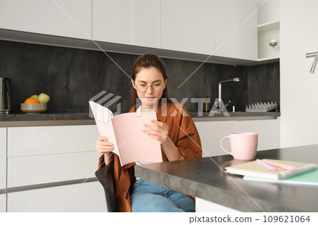 Portrait of young woman working from home, self-employed entrepreneur reading her documents, sitting at home in kitchen, wearing glasses Portrait of young woman working from home, self-employed entrepreneur reading her documents, sitting at home in kitchen, wearing glasses 109621064
