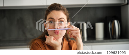 Close up portrait of funny, silly young woman, playing with pen, holding pencil on top of her lip and grimacing, sitting in kitchen 109621093