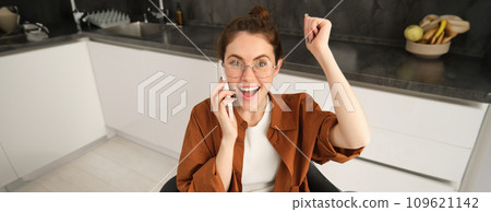 Portrait of woman receiving great news over the phone. Girl talking on mobile telephone and celebrating, laughing and making fist pump, dancing on her chair in kitchen 109621142