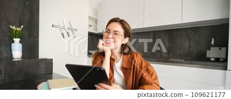 E-learning and remote workplace concept. Young woman in glasses, student studying at home in kitchen, reading on digital tablet, looking at her project, connects to online meeting 109621177