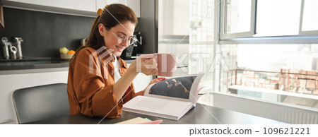 Portrait of woman reading at home, flipping pages of favourite book, relaxing in kitchen with cup of coffee, wearing glasses 109621221