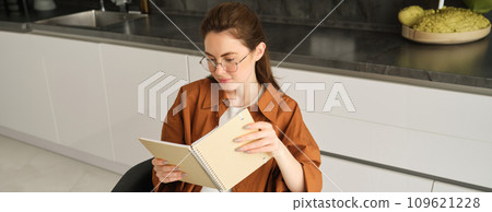 Close up portrait of young woman, student in kitchen, holding notebook, revising for exam at home, studying, reading her planner Close up portrait of young woman, student in kitchen, holding notebook, revising for exam at home, studying, reading her planner 109621228