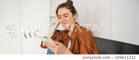 Young woman sitting at home on kitchen counter and using her smartphone. Concept of social media, leisure and mobile connection Young woman sitting at home on kitchen counter and using her smartphone. Concept of social media, leisure and mobile connection 109621232