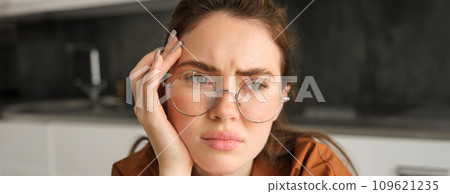 Close up portrait of woman in glasses with concerned, sad face, looking upset, troubled expression, sitting at home 109621235