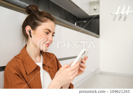 Close up portrait of young modern woman sits on kitchen floor, looking at her smartphone, watching video or tv series on streaming service app, listening music in wireless headphones Close up portrait of young modern woman sits on kitchen floor, looking at her smartphone, watching video or tv series on streaming service app, listening music in wireless headphones 109621265