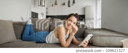 Portrait of cute, happy young woman lying on couch, holding mobile phone, using smartphone while relaxing at home in living room, concept of lifestyle and people leisure 109621327
