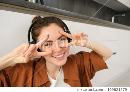 Close up of carefree young modern woman, relaxes at home, listens music in wireless headphones, shows v-sign and smiles at camera Close up of carefree young modern woman, relaxes at home, listens music in wireless headphones, shows v-sign and smiles at camera 109621379