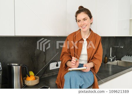 Portrait of stylish young woman, sits at home in the kitchen and makes notes, writes down recipe, checks her list with house errands, doodling or drawing in notebook Portrait of stylish young woman, sits at home in the kitchen and makes notes, writes down recipe, checks her list with house errands, doodling or drawing in notebook 109621389