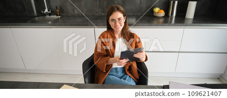 Portrait of young self-employed woman sitting at home in kitchen, reading papers, holding digital tablet, wearing glasses Portrait of young self-employed woman sitting at home in kitchen, reading papers, holding digital tablet, wearing glasses 109621407