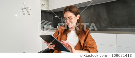 Young woman at home, reading in glasses on digital tablet and smiling, sitting in kitchen 109621418