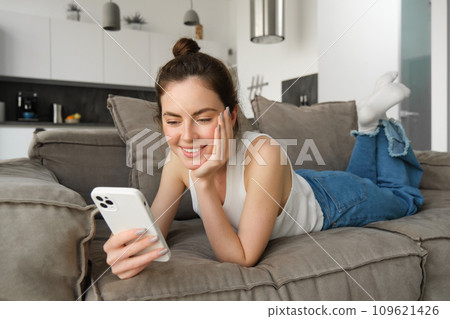 Portrait of smiling, happy young woman resting at home, resting on couch with smartphone, using mobile phone application Portrait of smiling, happy young woman resting at home, resting on couch with smartphone, using mobile phone application 109621426