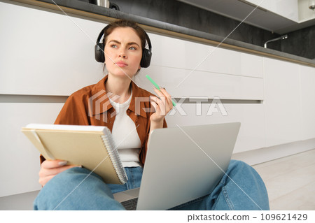 Portrait of young woman, modern student sits at home on floor and doing homework on laptop, listens music in headphones while making assignment or project for college Portrait of young woman, modern student sits at home on floor and doing homework on laptop, listens music in headphones while making assignment or project for college 109621429