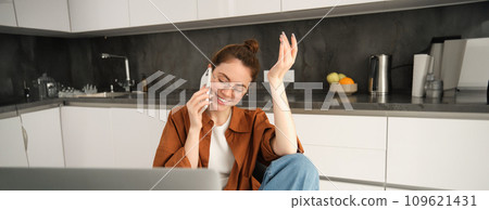 Excited young woman talking on mobile phone in front of laptop, sitting in kitchen with happy face expression, having a conversation Excited young woman talking on mobile phone in front of laptop, sitting in kitchen with happy face expression, having a conversation 109621431