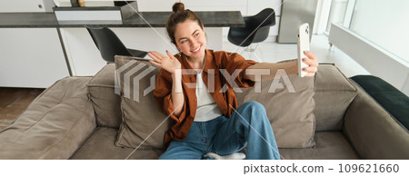 Photo of smiling young woman sitting on couch, posing on sofa in living room, taking selfies for social media 109621660