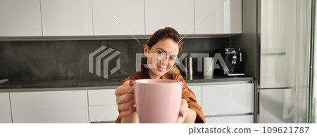 Beautiful young woman in glasses, giving you cup of tea, passing mug with aromatic delicious coffee, sitting in kitchen at home 109621787