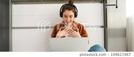 Woman looks surprised and amazed at laptop screen, hears shocking news on video chat, sits on floor at home 109621987