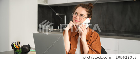 Portrait of young self-employed woman, entrepreneur working from home, freelancer calling client. Girl making an order, talking to someone on phone, sitting in kitchen with laptop Portrait of young self-employed woman, entrepreneur working from home, freelancer calling client. Girl making an order, talking to someone on phone, sitting in kitchen with laptop 109622015