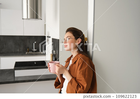 Portrait of happy young woman at home, holding cup of tea, leaning on wall, smiling, drinking coffee from pink mug 109622123