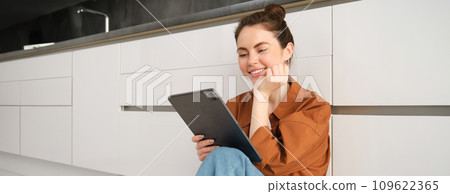 Portrait of beautiful, carefree young woman, sitting on kitchen floor with digital tablet, working remote from home, using device to check her mail. Girl watching something on her device 109622365