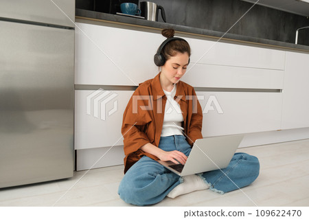 Young woman freelancer, working from home, student sitting with laptop on kitchen floor, wearing headphones, typing on computer 109622470