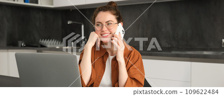 Portrait of beautiful smiling woman working from home, talking on mobile phone, calling client, self-employed businesswoman sets up workplace in kitchen, using laptop and smartphone 109622484