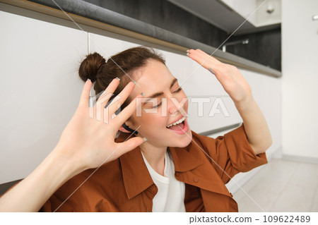 Carefree woman singing along song, listens to music in wireless headphones, enjoying sound quality, dancing with hands 109622489