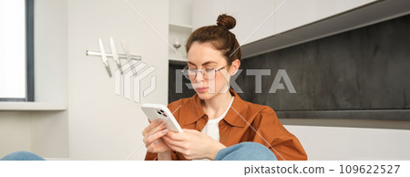 Portrait of young brunette woman in glasses, sitting at home with mobile phone, using her smartphone and smiling, reading message 109622527