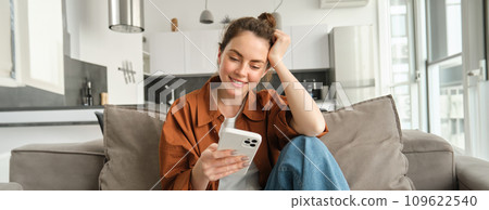 Portrait of happy young brunette woman sitting on sofa with smartphone, reading notification, messaging on mobile phone, resting on couch at home, smiling while looking at device screen 109622540
