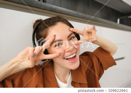Happy female model listens music, posing in wireless headphones, smiling and laughing, showing v-sign 109622661