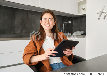 E-learning and remote workplace concept. Young woman in glasses, student studying at home in kitchen, reading on digital tablet, looking at her project, connects to online meeting 109622691
