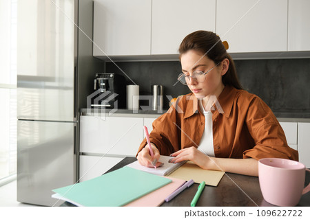 Portrait of woman looking concentrated, writing down notes, doing homework in kitchen, drinking coffee, studying at home 109622722