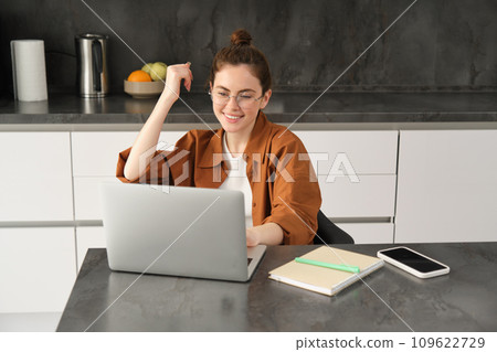 Portrait of young woman, business owner in glasses, sitting with laptop in kitchen, working from home. Student studying, doing homework on computer 109622729