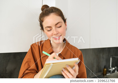Portrait of cute young woman with notebook, sitting in the kitchen and writing her to do list, making tasks for week, puts notes in schedule or planner Portrait of cute young woman with notebook, sitting in the kitchen and writing her to do list, making tasks for week, puts notes in schedule or planner 109622766