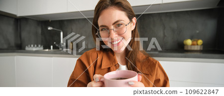 Close up portrait of beautiful woman in glasses, drinks tea, warms her hands with hot cup, smiling and looking happy at camera, sitting in kitchen 109622847