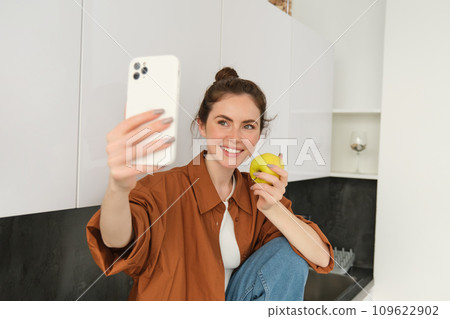 Portrait of happy, smiling young woman records herself, takes selfie while eating an apple in the kitchen, using smartphone app, makes photos with mobile phone 109622902