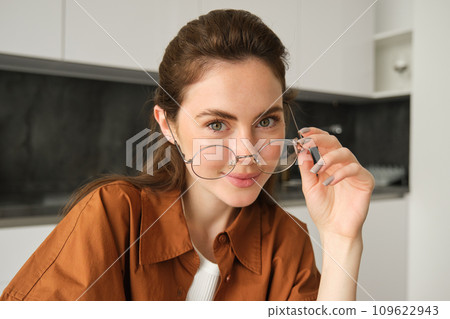 Portrait of beautiful young woman, smiling and looking happy, takes-off her glasses and looking at camera, sitting in kitchen at home 109622943
