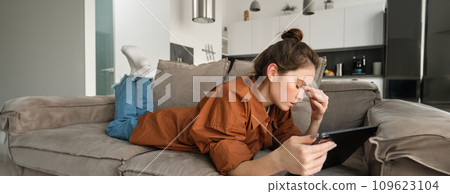 Portrait of woman resting on couch at home, rubs her eyes after reading too long on digital tablet, feeling tired, spending time in living room on sofa 109623104