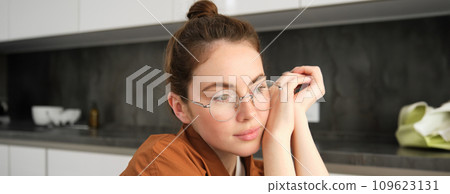 Close up portrait of gorgeous young woman in glasses, looking thoughtful and relaxed, posing sitting in kitchen, smiling with tenderness Close up portrait of gorgeous young woman in glasses, looking thoughtful and relaxed, posing sitting in kitchen, smiling with tenderness 109623131