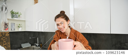 Portrait of cute girlfriend offering a cup of coffee, extending hand with mug and smiling, sharing her drink with someone, sitting in kitchen Portrait of cute girlfriend offering a cup of coffee, extending hand with mug and smiling, sharing her drink with someone, sitting in kitchen 109623146