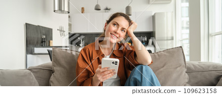 Smiling brunette woman sitting on sofa with smartphone, looking thoughtful, thinking while using mobile phone app, ordering on application, doing online shopping and resting at home 109623158
