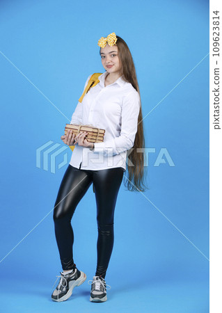 Charming female student with long hair carrying bunch of books, while smiling at camera. Side view of pretty teen girl in shirt standing, shyly bending one leg, isolated on blue. Concept of studying. 109623814