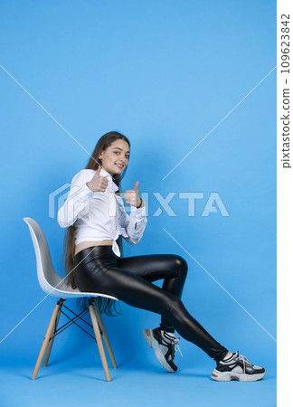 Cheerful caucasian lady keeping thumb up, while resting in white cozy chair. Side view of smiling woman showing okay sign at camera, while leaning back, isolated on blue. Concept of approval gesture. 109623842