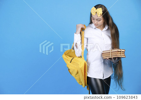 Young female with very long hair looking back, while wearing yellow backpack. Front view of cute brunette with bunch of books prepare to study, isolated on blue background. Concept of studying.  109623843