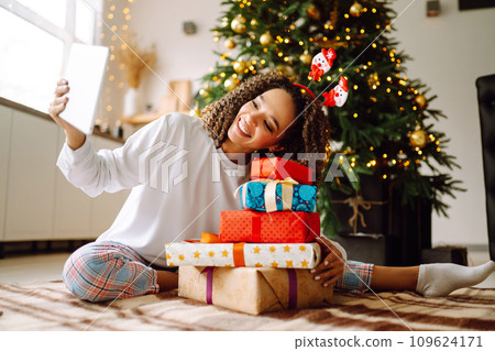 Young woman on background of Christmas tree with gifts, with tablet has video call or video chat. Young woman on background of Christmas tree with gifts, with tablet has video call or video chat. 109624171