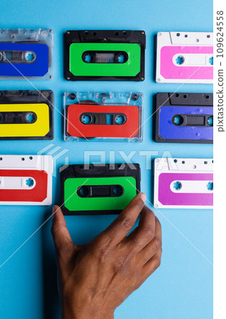 Overhead view of african american hand with colourful cassette tapes arranged on blue background Overhead view of african american hand with colourful cassette tapes arranged on blue background 109624558